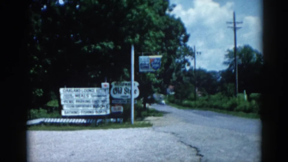 8mm home-movie frame from c.1962 Illinois showing roadside signs advertising the Oakland Lounge Resort, listing meals, rooms, fishing boats, and a pool, alongside an Old Style beer sign.