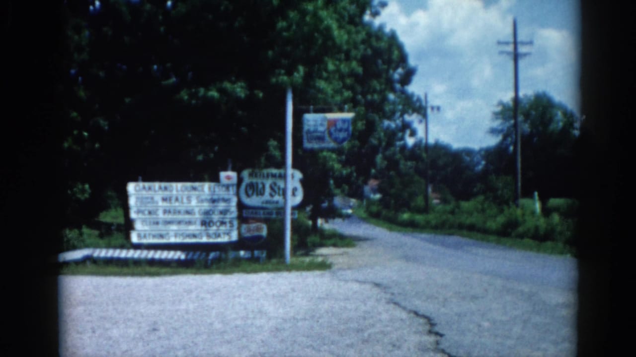 Second 8mm frame from c.1962 Illinois showing the same roadside resort sign cluster from a similar angle, with dense summer tree canopy overhead.