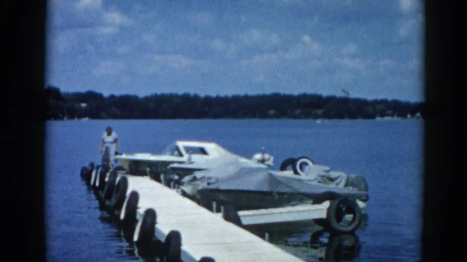 8mm frame from c.1962 Illinois showing a man on a floating dock next to a canvas-covered boat, with calm lake water and a wooded shoreline behind him.