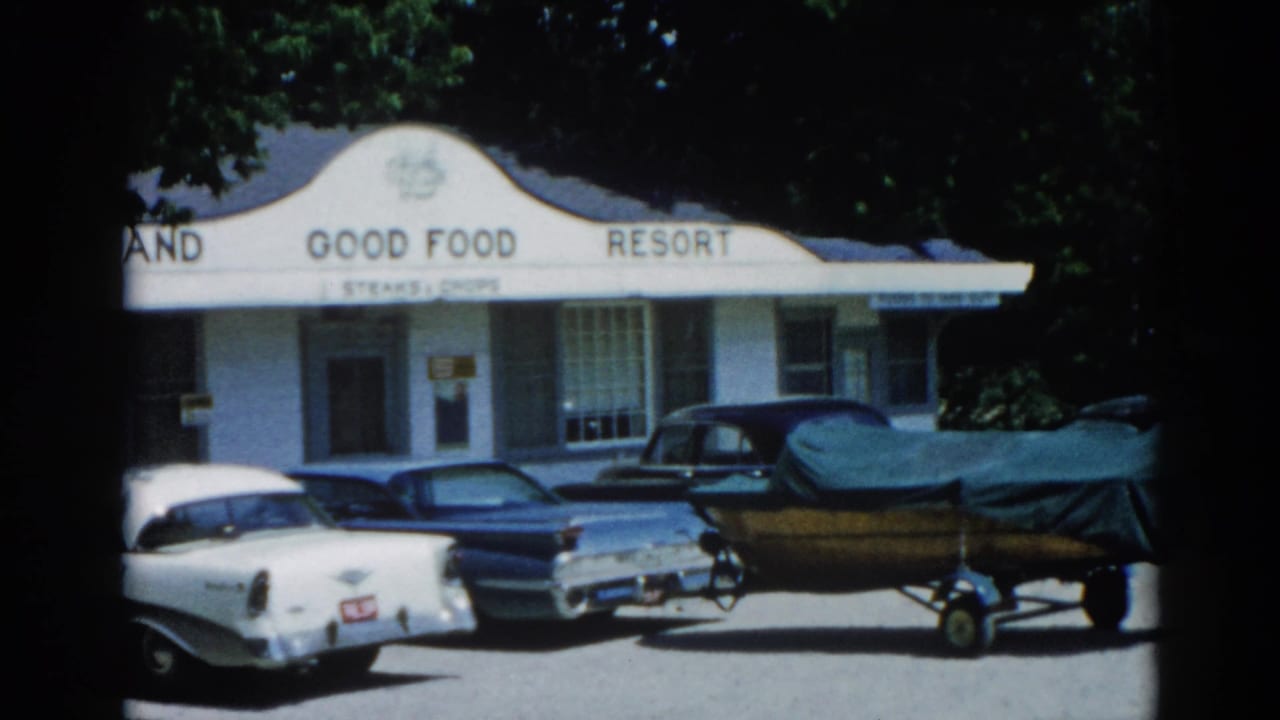 White 'GOOD FOOD RESORT' building with fin-era American cars and a covered boat on trailer parked in front, leafy trees above.