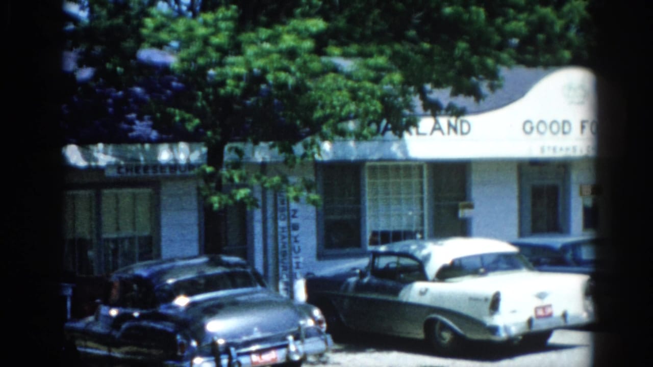 White arched building facade partially reading 'Oakland Good Food' with vintage cars parked below, framed by green tree canopy.