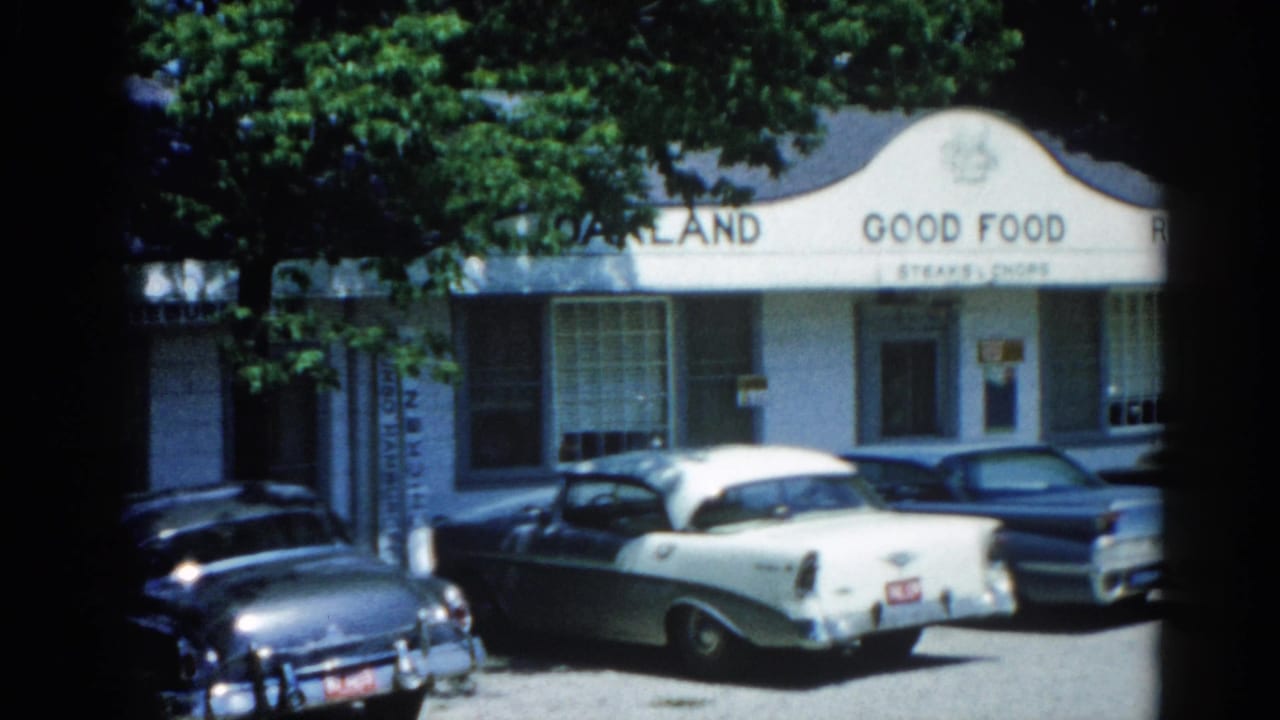 White arched commercial building with 'Oakland Good Food' visible on facade and multiple vintage sedans in the parking lot.