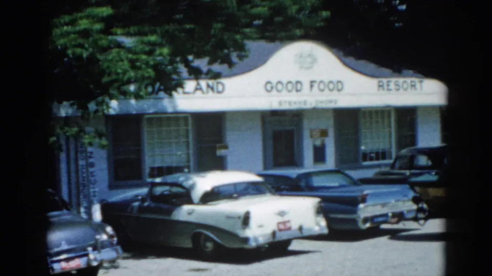 8mm home movie frame circa 1962, Illinois: complete 'Oakland Good Food Resort' signage with a secondary menu line below, late-1950s sedans in foreground.
