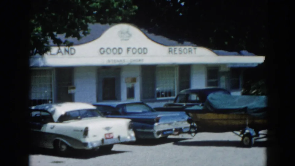 8mm home movie frame circa 1962, Illinois: 'Oakland Good Food Resort' with late-1950s sedans and a blue boat on a trailer at right, suggesting a nearby waterway.