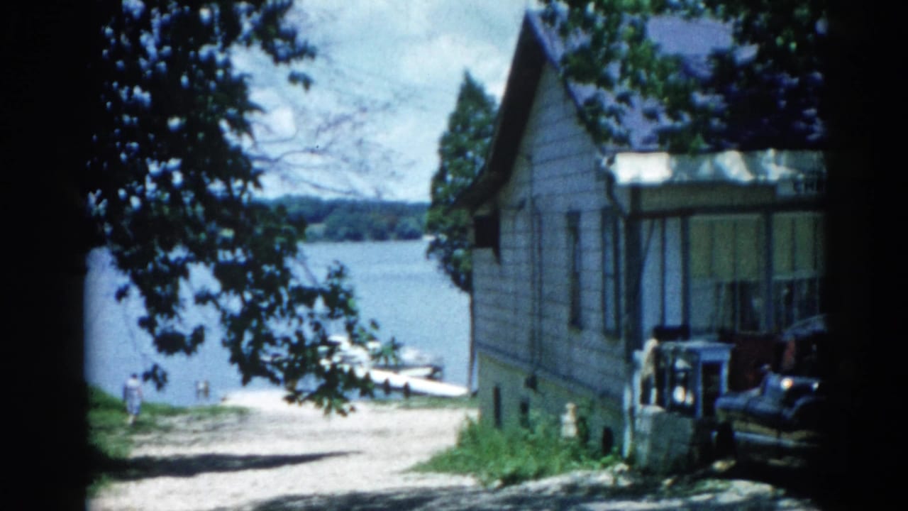 Gray clapboard lakeside house with a large tree, blue lake behind, and a dark early-1960s car partially visible at right.