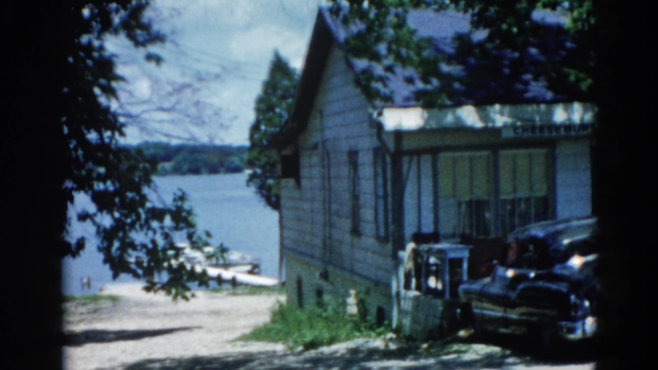 Second 8mm frame from 1962 Illinois showing the same lakeside house and grounds with a dark period automobile parked alongside.
