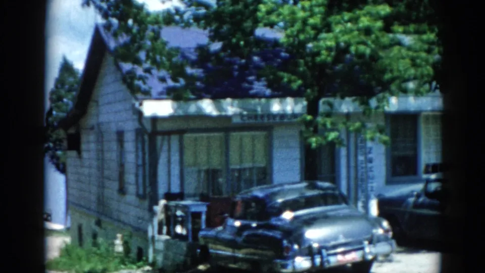 8mm frame circa 1962 Illinois: multiple early-1960s American sedans parked before a gray structure with partially visible commercial signage.