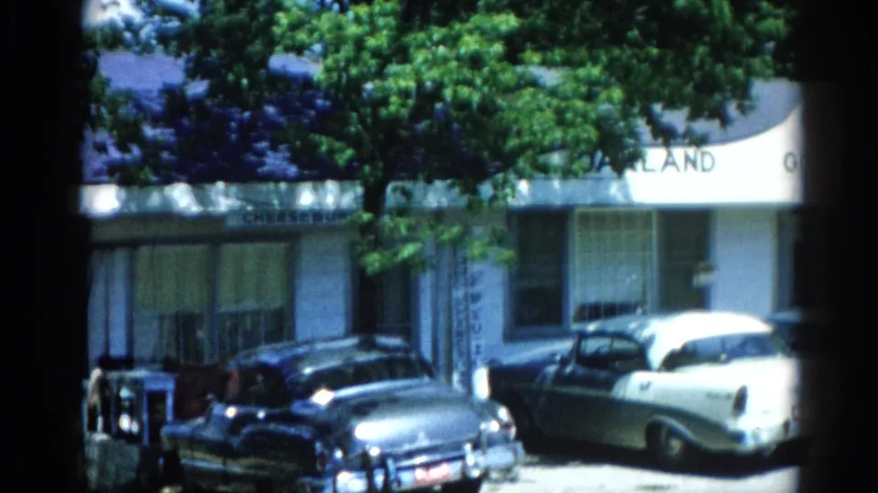 8mm frame circa 1962 Illinois: three period American automobiles parked outside a gray clapboard building, with partially legible signage visible.
