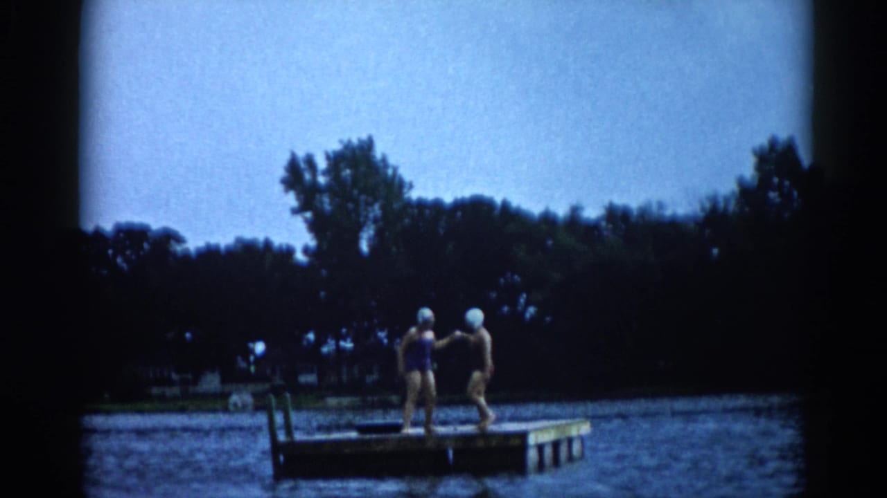 Two young women in early-1960s one-piece swimsuits and white rubber swim caps stand on a floating dock at an Illinois lake, c. 1962.
