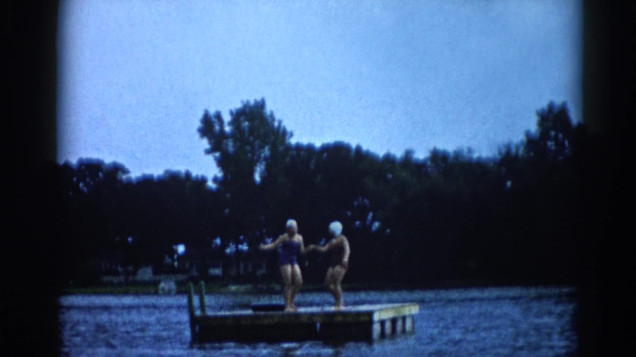 Two young women in white rubber swim caps and dark swimsuits turn toward each other on a floating dock, Illinois lake, c. 1962.