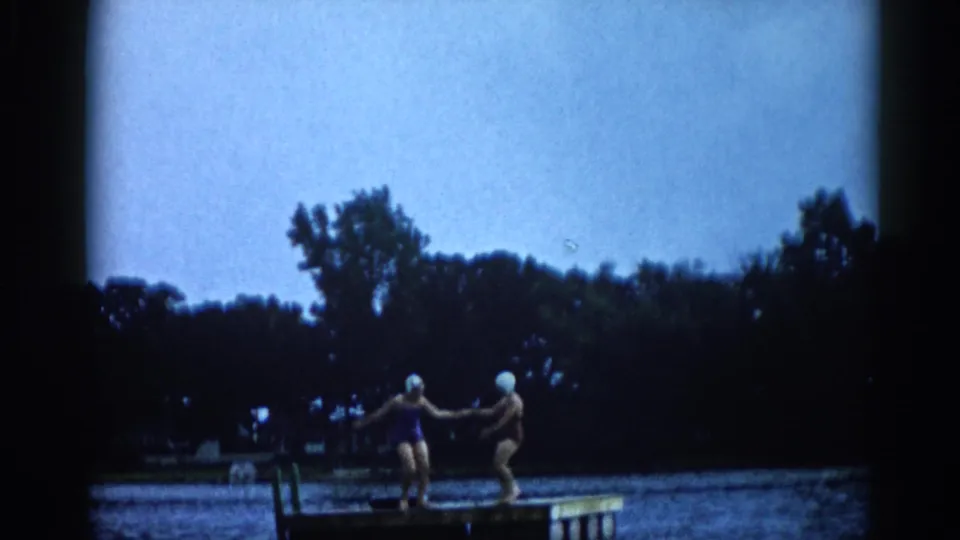 Two young women in 1960s swimwear reach toward each other on a lake dock in Illinois, c. 1962, with a dense treeline behind them.