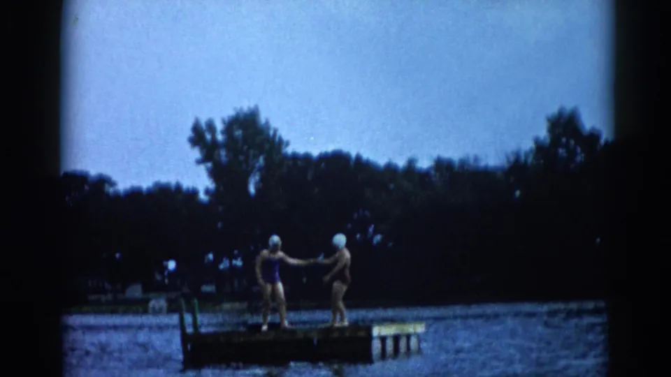Two young women in 1960s swimsuits and white rubber swim caps hold hands on a floating lake dock, Illinois, c. 1962.