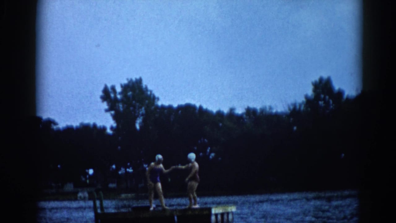 Two women in white swim caps and dark bathing suits standing on a wooden lake dock, facing each other.