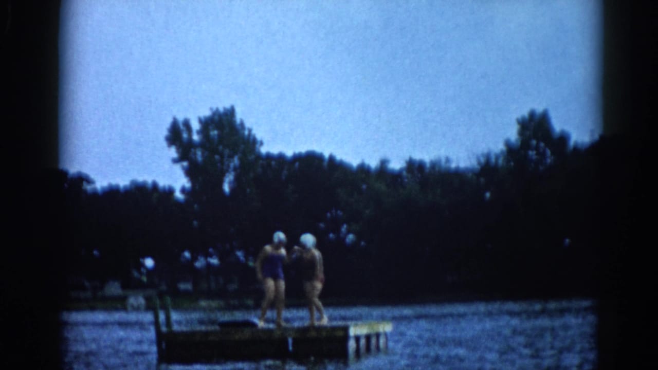Two women in vintage swim caps stand together on a dock over an Illinois lake, with white globe park lamps visible in the mid-background, circa 1962.