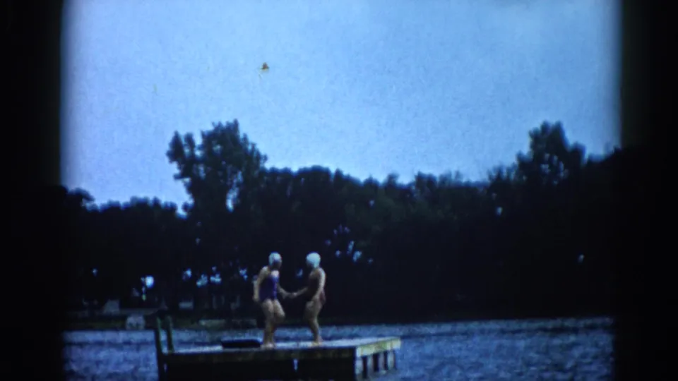Two women in white swim caps and dark bathing suits dance on a lake dock in Illinois, arms extended, circa 1962, framed at wider distance.