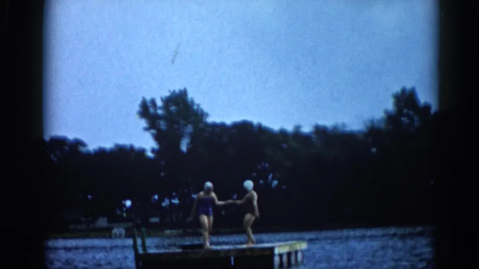 Two women in white swim caps perform a joyful dance on a lake dock in Illinois, one with a leg raised, circa 1962.