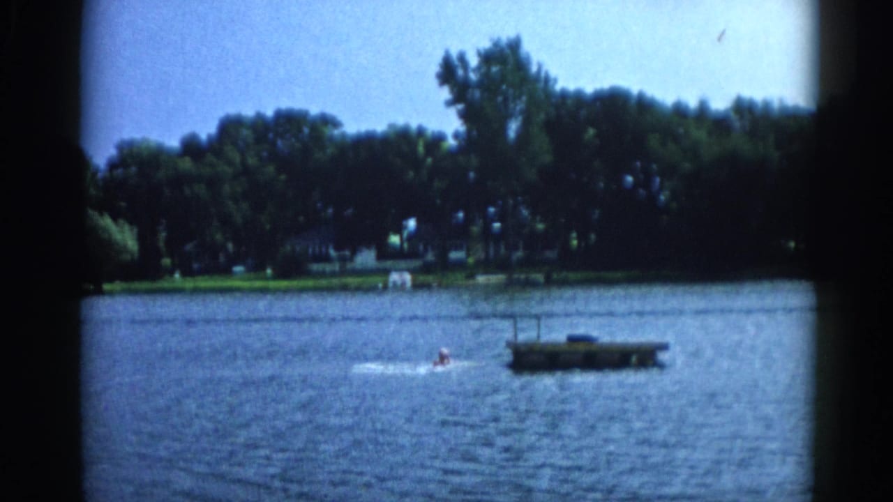 Two swimmers in a lake approaching a square wooden floating dock, with a dense tree-lined shore in the background.