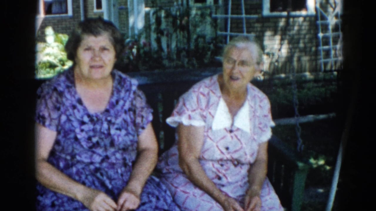 Two adult women, likely family members, seated together in a sunlit backyard in Illinois, c. 1962; 8mm home-movie frame with characteristic film grain.