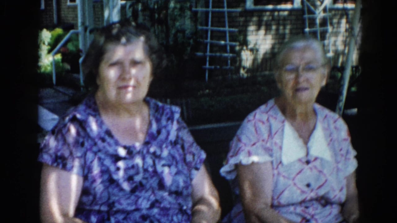 Two adult women seated together in a sunlit backyard, Illinois, c. 1962; mid-shot from an 8mm home-movie reel with warm period color.
