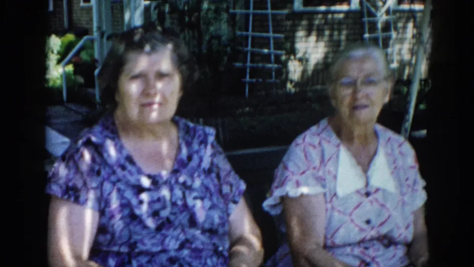 Two adult women seated together in a sunlit backyard, Illinois, c. 1962; mid-shot from an 8mm home-movie reel with warm period color.