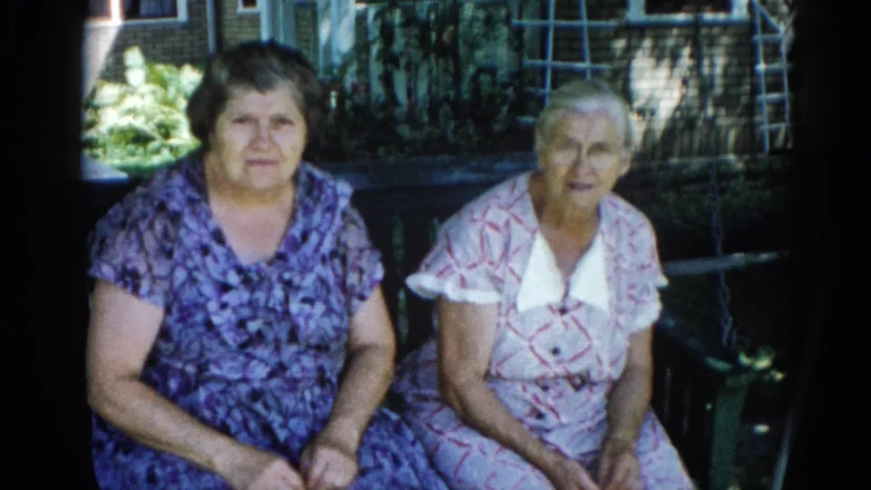 Two adult women seated together in a sunlit backyard, Illinois, c. 1962; 8mm home-movie footage with consistent static framing.