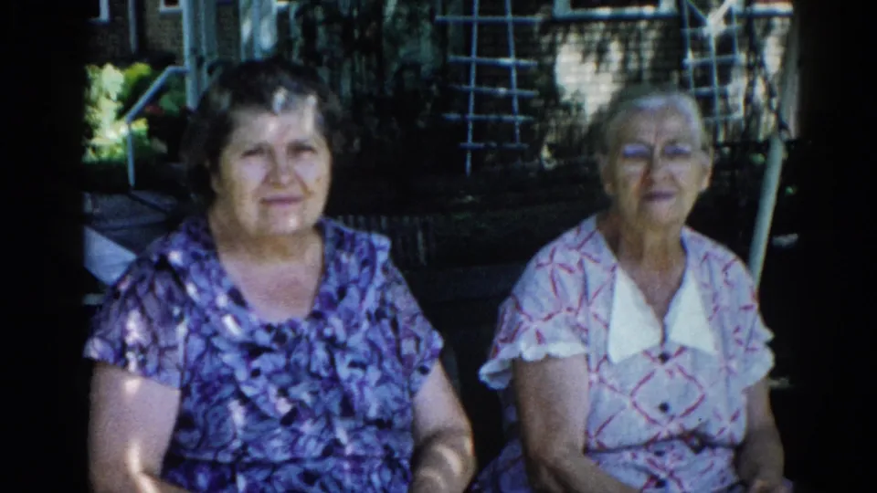 Two adult women in a sunlit backyard, younger woman smiling toward camera; 8mm home-movie frame, Illinois, c. 1962.