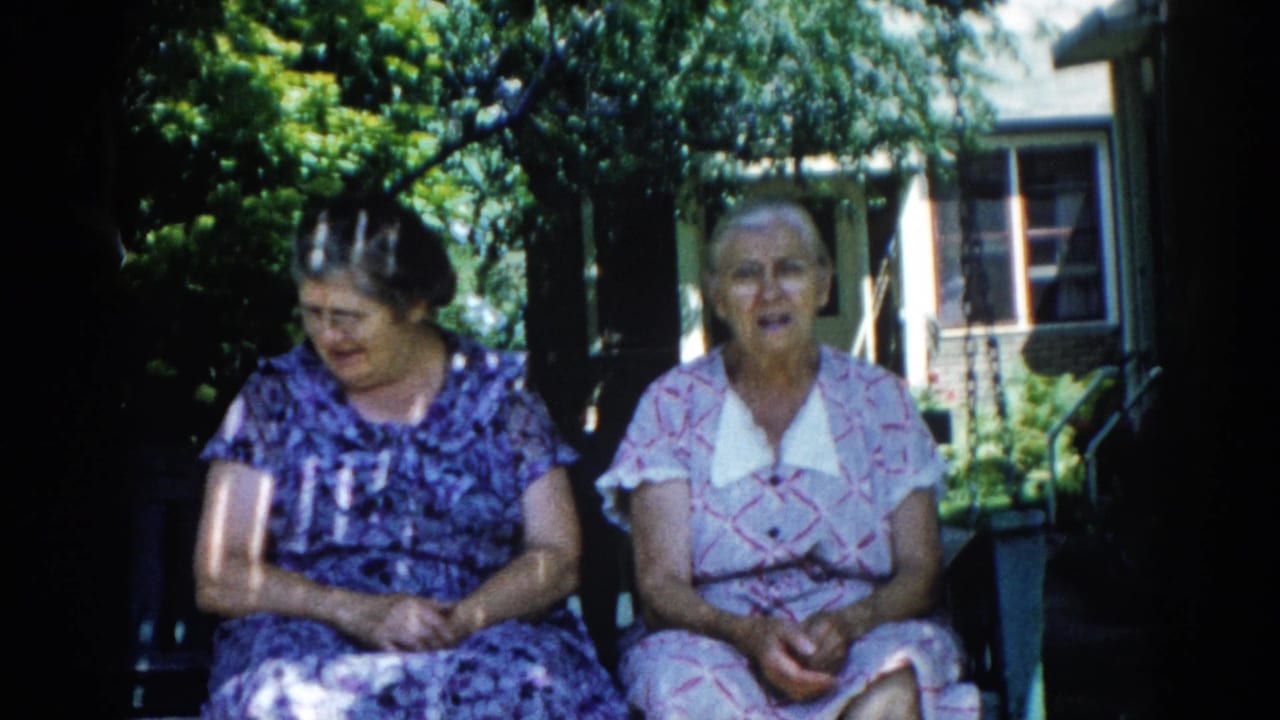 Two older women in early-1960s patterned house dresses seated on a wooden porch swing outside a brick house, Illinois, 1962.