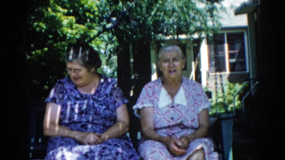 Two older women in early-1960s patterned house dresses seated on a wooden porch swing outside a brick house, Illinois, 1962.