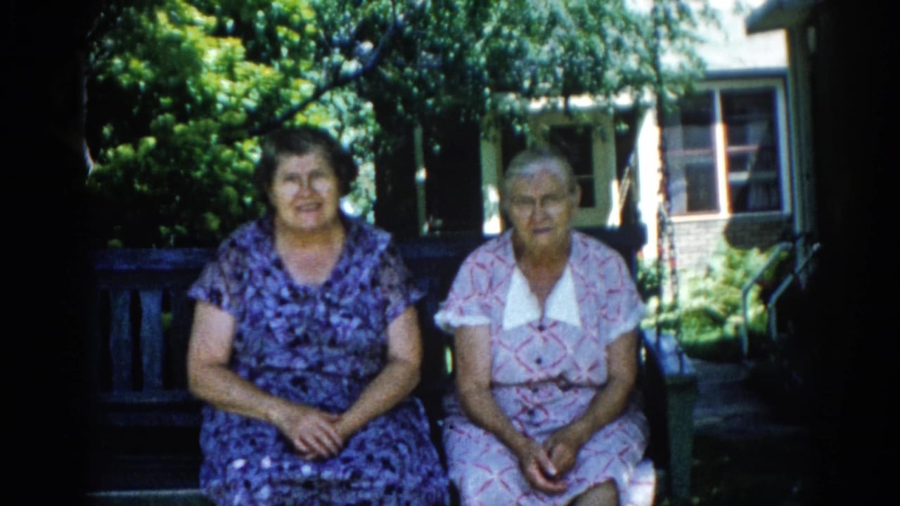 Two older women on a porch swing; woman on left turns toward woman on right; wooden lattice and green foliage visible behind them.