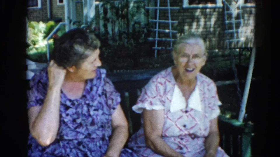 Two older women in animated conversation on a porch swing, Illinois, 1962; woman on left raises hand to her face mid-speech, mouth open.