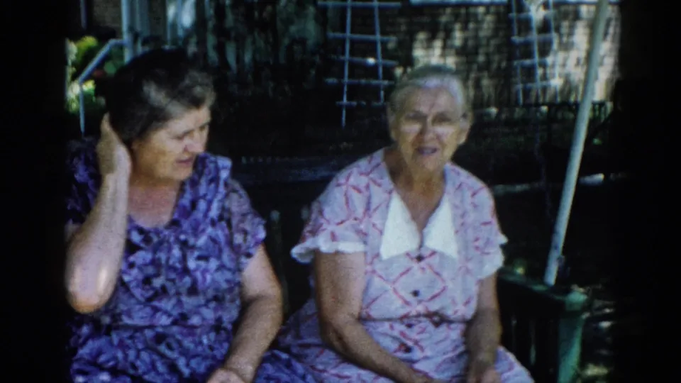 Two older women on a porch swing in Illinois, 1962; woman on left holds hand to side of head; wooden lattice and green foliage visible behind.