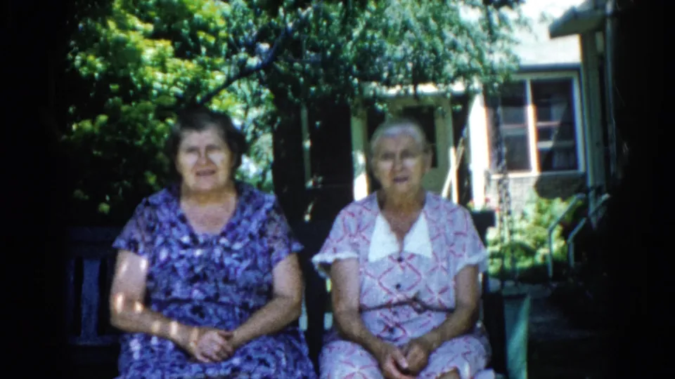 Continued 8mm shot of two women on a porch swing, Illinois, 1962 — both in patterned dresses with house exterior and green garden shrubs in the background.