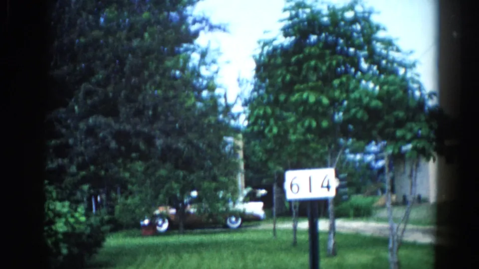 Continuation of the Illinois suburban establishing shot, circa 1962, with address marker '614' and classic car visible under full tree canopy.