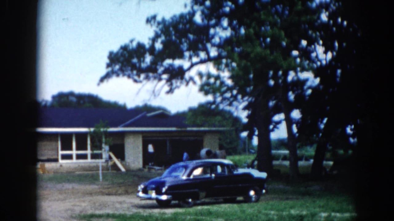 Dark early-1950s sedan parked beside a single-story ranch house under large trees, Illinois 8mm home movie.