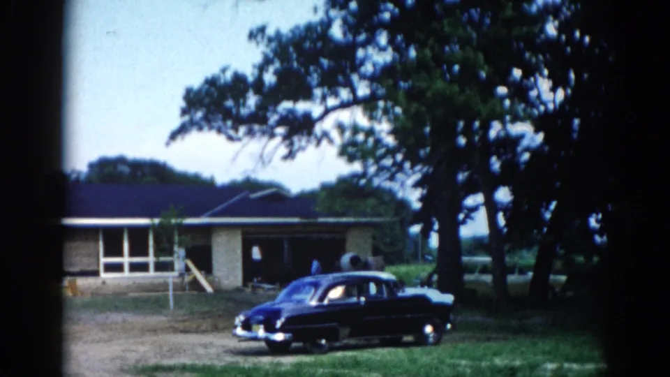 Early-1950s dark-colored American sedan parked on dirt in front of a single-story ranch house with dark roof, mature trees, and green lawn; 8mm home movie, Illinois, 1962.