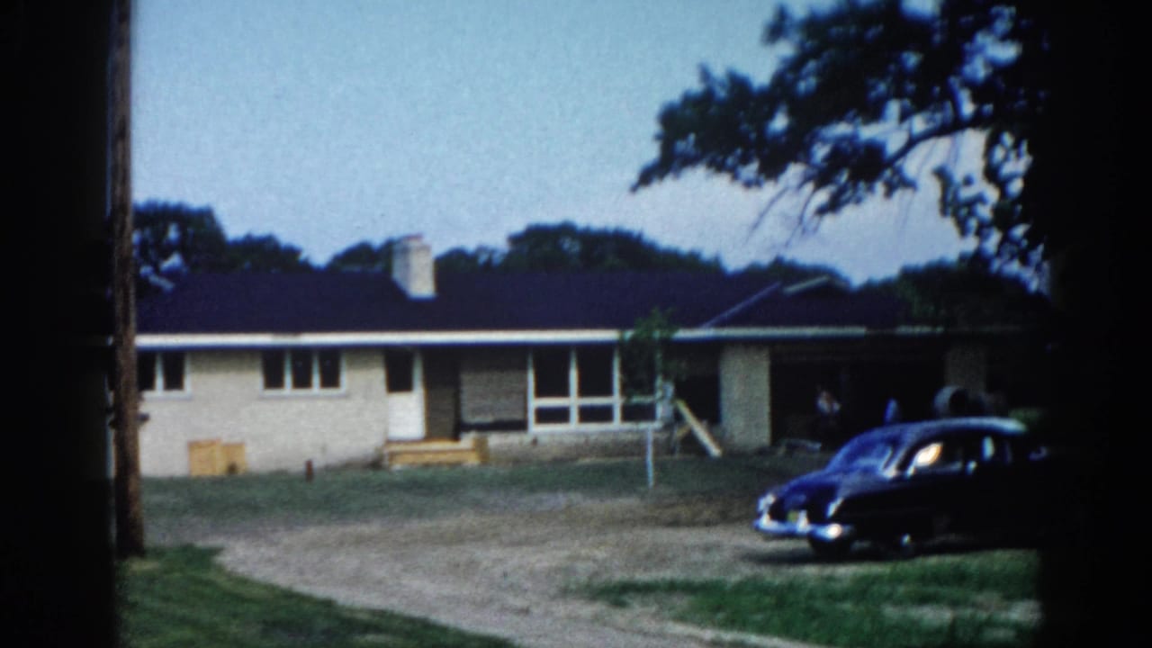 Dark classic sedan parked on gravel driveway before a single-story ranch-style home; large trees and open sky in background.