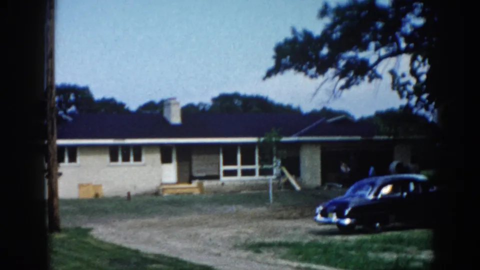 Dark American sedan, likely late-1940s to early-1950s vintage, parked outside a ranch-style home in Illinois circa 1962, captured on 8mm film with characteristic warm grain.