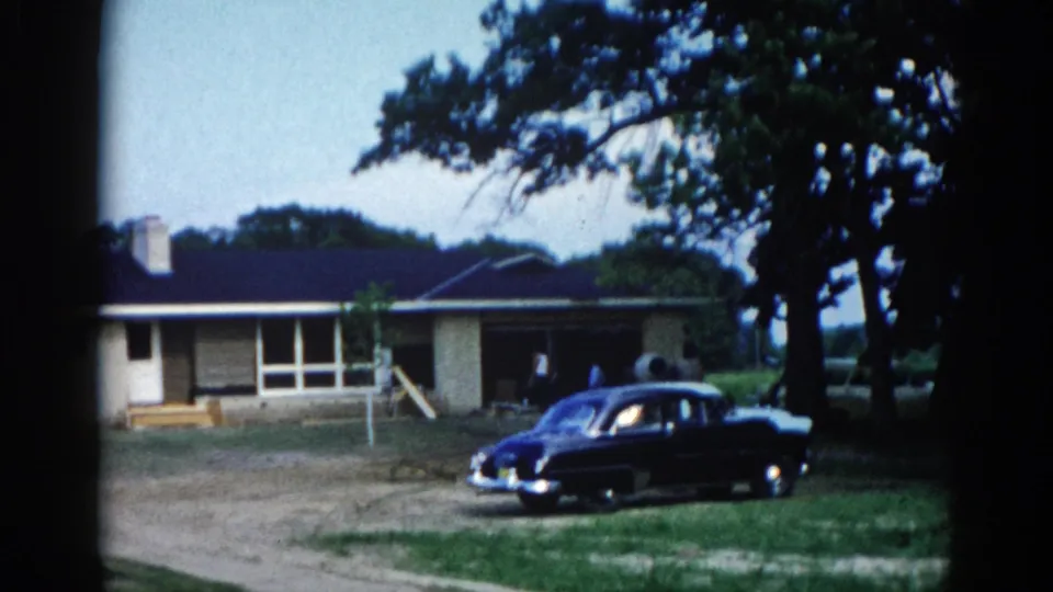 8mm frame of ranch-style home in Illinois circa 1962 with dark sedan on driveway; camera may have shifted slightly, foregrounding the mature deciduous tree on the right.