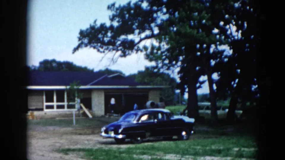 Closing 8mm frame of ranch-style home and dark American sedan in Illinois circa 1962; static hold with mature trees and open sky visible, consistent 8mm film grain throughout.