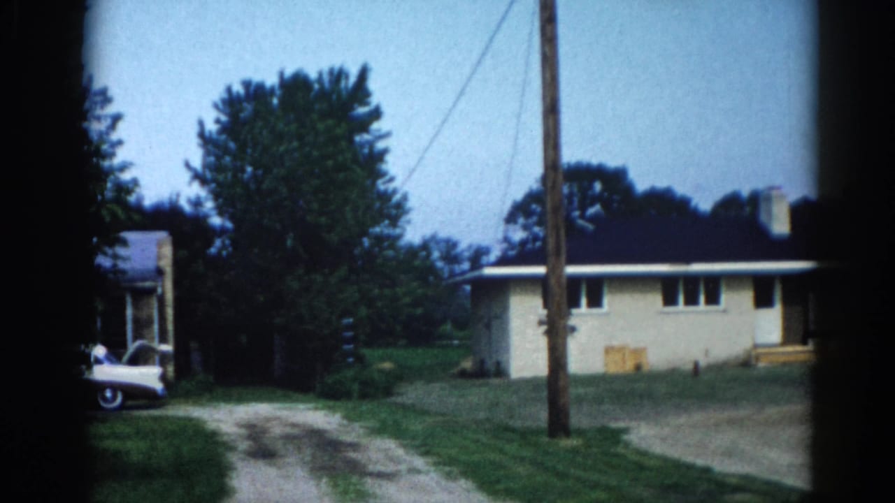 8mm home movie frame, c.1962, Illinois — cream ranch house with dark roof, gravel driveway, utility pole with power lines, white car at left edge, mature trees, blue sky.