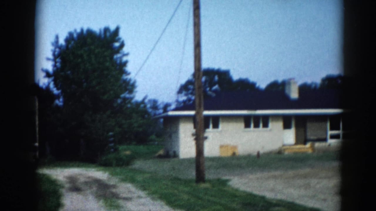 8mm home movie frame, c.1962, Illinois — cream single-story ranch house, utility pole with power lines, yellow boxes at foundation, gravel foreground, summer trees, blue sky.