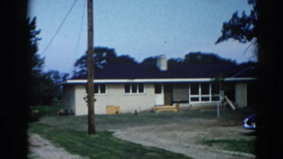 8mm home movie frame, c.1962, Illinois — cream ranch house with front entrance steps, dark blue classic American automobile entering bottom of frame, utility pole, mature trees.