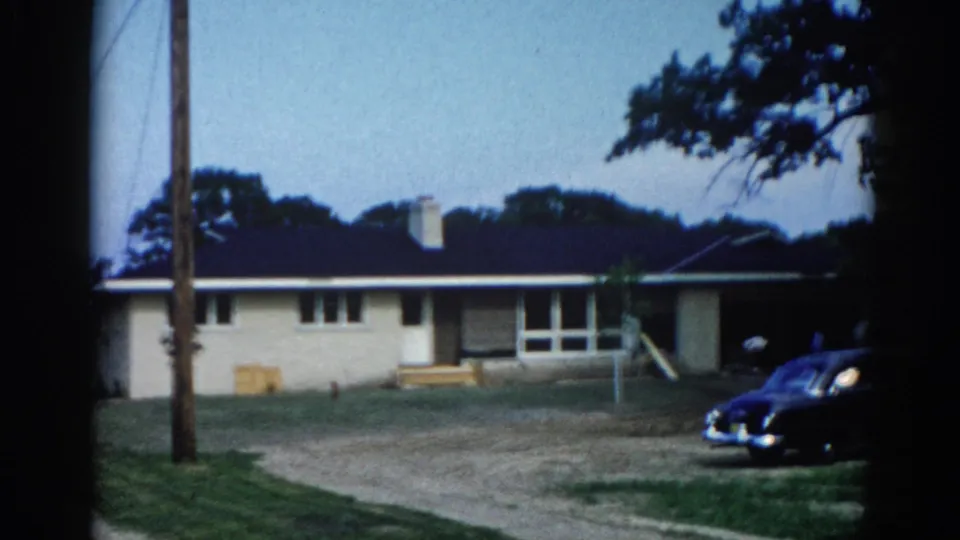 8mm home movie frame, c.1962, Illinois — cream ranch house, dark blue late-1950s/early-1960s automobile parked at right, utility pole, gravel lot, mature trees, blue sky.