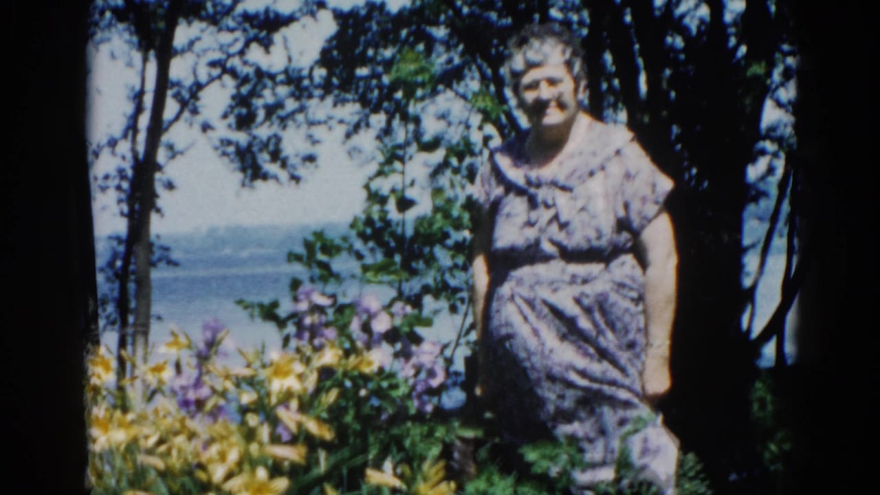 Middle-aged woman posing in a garden amid yellow and purple flowers, water visible behind; 1962 Illinois 8mm home movie.