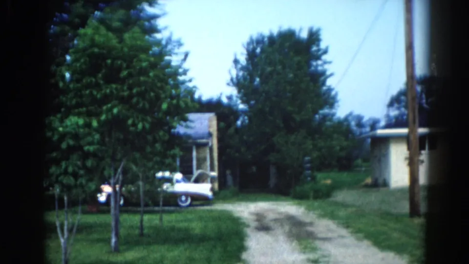 Ranch-style house with light-colored siding, white automobile on gravel driveway, and utility poles; consistent with early-1960s Midwest suburban setting.