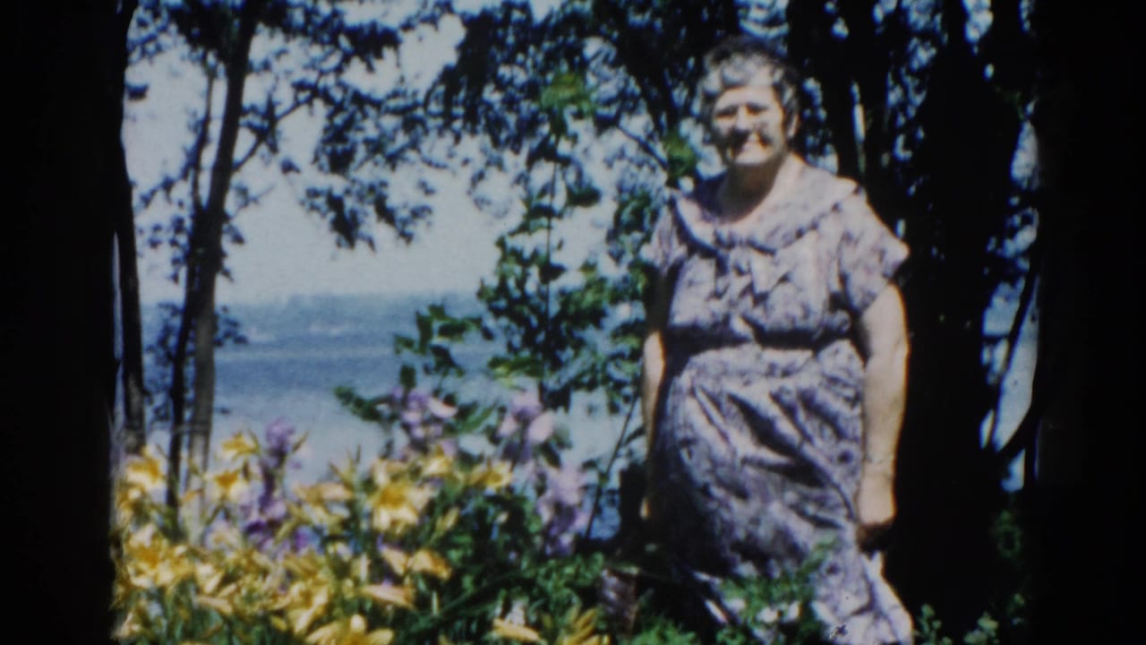 Elderly woman in patterned dress standing in a garden with yellow daylilies and purple flowers, lake visible behind dark tree trunks.
