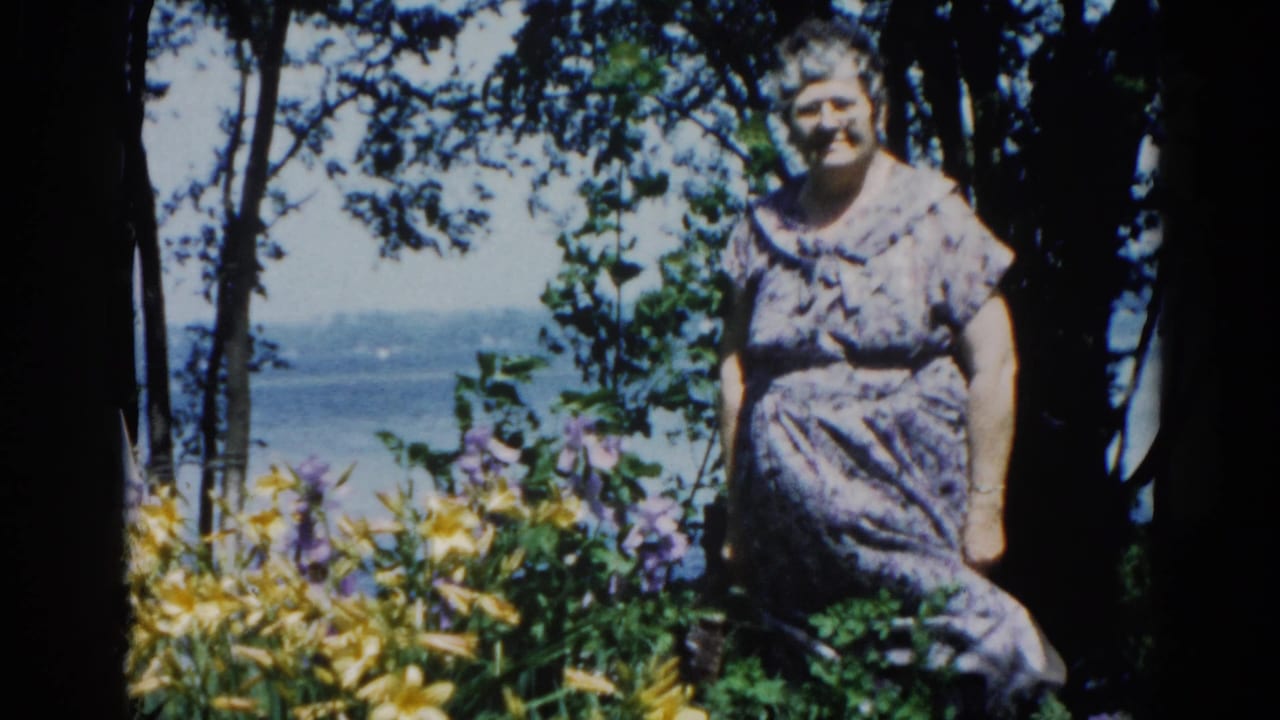 Elderly woman in patterned dress standing amid garden flowers with dark trees and water behind her.