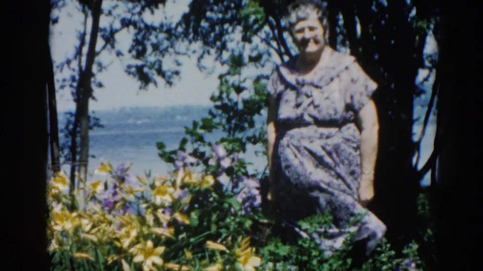 Elderly woman in floral housedress in windswept lakeside garden, yellow daylilies prominent, Illinois circa 1962, filmed on 8mm.