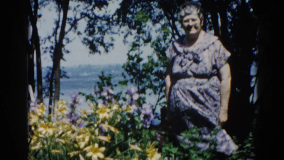 Medium close shot of a smiling woman in a print dress in an Illinois lakeside garden, circa 1962; yellow and purple flowers flank her on both sides.