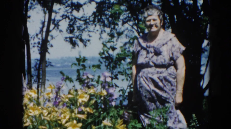 Woman in a floral-print dress poses in an Illinois lakeside garden, circa 1962; yellow daylilies and tall trees frame the scene behind her.